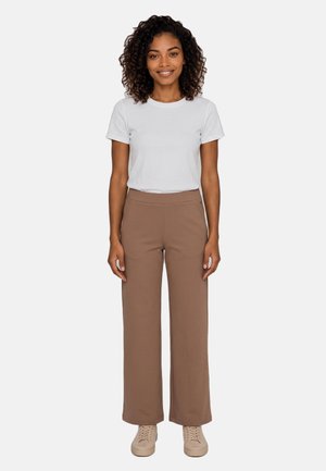 Mujer con pelo rizado está de pie sonriendo, lleva una camiseta blanca de manga corta, pantalones marrones de pierna ancha y zapatillas beige sobre un fondo blanco.
