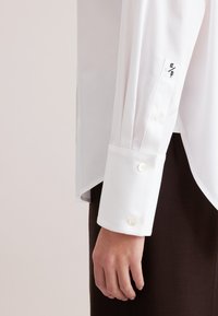 Camicia bianca con bottoni, maniche lunghe, dettagli plissettati e polsini con bottone. Il tessuto appare liscio e crispato, con una vestibilità sfiancata.