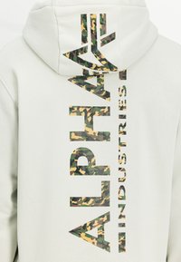 Ανοιχτό γκρι φούτερ με κουκούλα που διαθέτει ένα μεγάλο, κάθετο λογότυπο με καμουφλάζ και την επιγραφή "ALPHA INDUSTRIES" σε στυλιζαρισμένα, μπλοκ γράμματα.