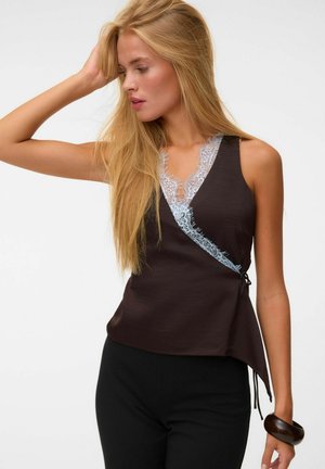 Femme aux longs cheveux blonds portant un haut sans manches marron foncé avec une bordure en dentelle bleu clair et un pantalon noir, posant avec une main sur la tête.