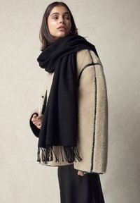 Cappotto teddy color crema con piping nero, abbinato a una lunga sciarpa nera con frange. Texture liscia e silhouette morbida.