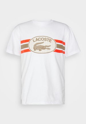 T-shirt en coton blanc à manches courtes et col rond. Présente un grand logo ovale avec un crocodile ainsi que des rayures rouges, marron et beiges.