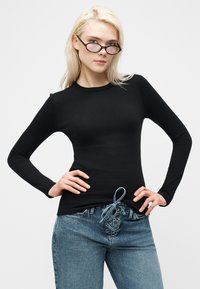 Νεαρή γυναίκα με μακριά ξανθά μαλλιά, φορώντας μαύρο εφαρμοστό μακρυμάνικο ριπ τοπ, μπλε τζιν με κορδόνια και σκούρα γυαλιά ηλίου, με τα χέρια στη μέση.