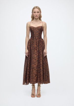 Γυναίκα στέκεται φορώντας ένα αμάνικο μίντι φόρεμα με καφέ και μαύρο λεοπάρ μοτίβο, λεπτές τιράντες και χρυσά ανοιχτά τακούνια, με φόντο λευκό.