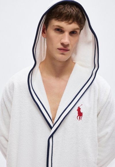 Νεαρός άνδρας που φοράει λευκή ρόμπα με κουκούλα και μπλε περιθώριο, καθώς και κόκκινο λογότυπο παίκτη πόλο, κοιτάζοντας μπροστά σε απλό φόντο.