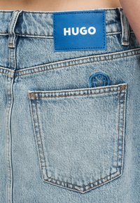 Κοντινό πλάνο πίσω τσέπης σε ανοιχτό μπλε τζιν με ραμμένη μπλε ετικέτα "HUGO" πάνω από την τσέπη.