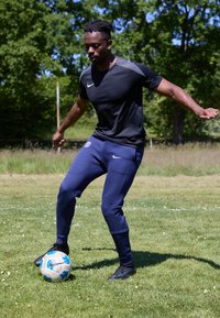 Nike must musta lühikeste varrukatega särk, tumesinised kitsenevad püksid, mustad sportlikud kingad, lööb sinise ja valge jalgpalli murul.