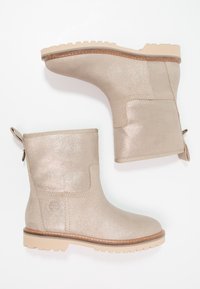 Beige třpytivé kotníkové boty s kulatou špičkou, texturovaným povrchem a béžovými gumovými podešvemi. Obsahují poutko pro snadné obouvání a logo detail po straně.