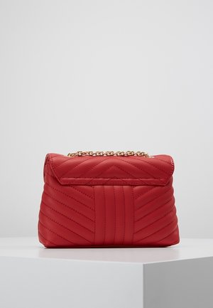 Rote, gesteppte Lederhandtasche mit Chevron-Steppung und goldenem Kettenriemen auf einem weißen, geometrischen Podest.