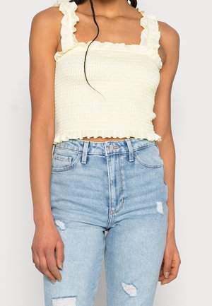 Frau trägt ein blassgelbes, gerafftes Crop-Top mit Rüschenträgern und hellblaue, hoch taillierte, zerrissene Jeans, steht vor einem schlichten Hintergrund.