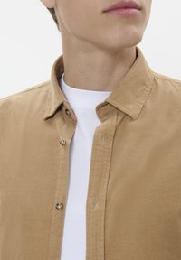 Beige corduroy button-up overhemd met een kraag, voorzien van goudkleurige knopen en een wit T-shirt met ronde hals eronder.