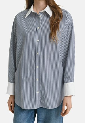 Kvinde iført en blå og hvid vertikalt stribet skjorte med hvid krave og manchetter, kombineret med blå jeans.