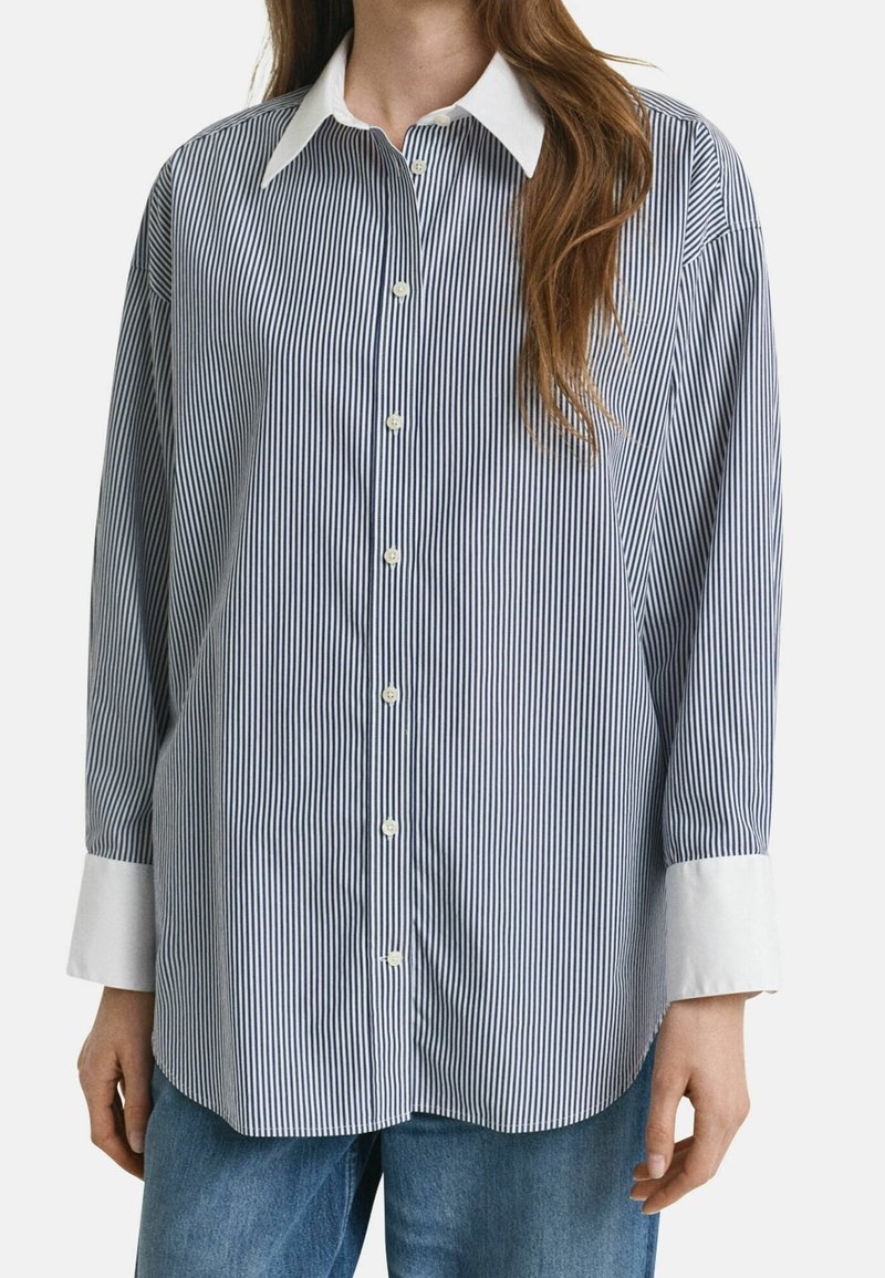 Femme portant une chemise à rayures verticales bleues et blanches avec col et poignets blancs, associée à un jean bleu.