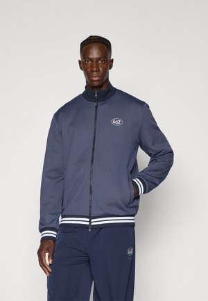 Marine blå zip-jakke med høy krage, hvite striper ved ermene og nederst, teksturert stoff og brodert logo på brystet.