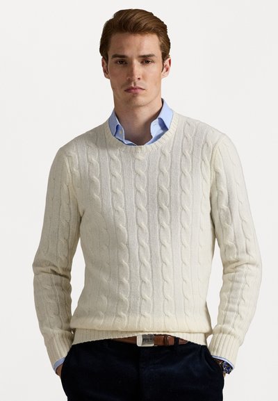 Un pull crème en maille torsadée avec un col rond et des poignets côtelés, porté sur une chemise à col bleu clair. Un pantalon sombre complète le look.