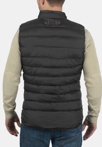 Schwarze Steppweste mit horizontalen Linien, Kragen und braunem Lederetikett. Getragen über einem hellgrauen Langarmshirt, mit blauen Jeans.