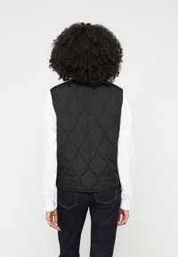 Asmuo su garbanotais plaukais, vilkintis juodą siūtu liemenę, uždėtą ant balto ilgomis rankovėmis marškinėlio ir tamsių džinsų, parodytas iš nugaros prieš neutralų foną.