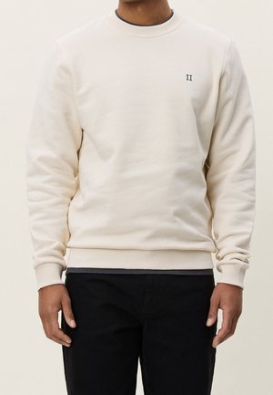 Homme portant un sweat-shirt col rond couleur crème avec un petit logo "II" sur la poitrine gauche et un pantalon noir, debout devant un fond uni.