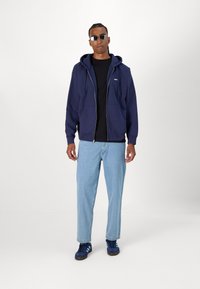 Navy zip-up hoodie with two pockets layered over a black t-shirt, paired with light blue jeans and navy shoes. Round sunglasses complete the look.