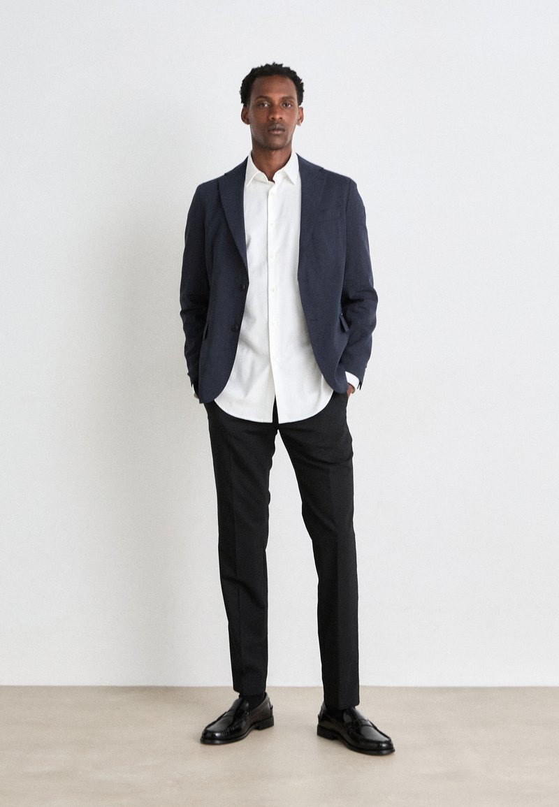 Blazer bleu marine sur une chemise blanche à boutons, associé à un pantalon noir et des chaussures de ville noires. Tissu lisse, coupe ajustée et design minimaliste.
