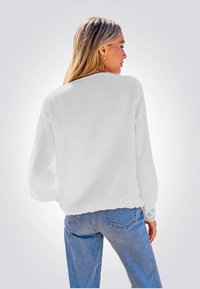 Witte, gestructureerde blouse met lange mouwen en ingewikkelde kantaccenten bij de manchetten, gecombineerd met lichtblauwe spijkerbroek. Achteraanzicht weergegeven.