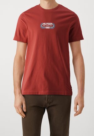 Mężczyzna w prostym czerwonym krótkim rękawem koszulce z napisem "Hilfiger New York City 1985" na klatce piersiowej i ciemnobrązowych spodniach.