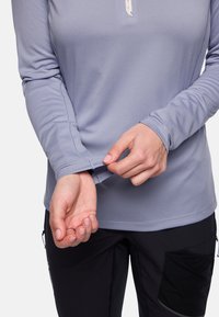 Hände, die den Kragen eines langärmeligen, hellvioletten Hemdes anpassen, das mit schwarzen Hosen mit Reißverschlusstaschen getragen wird.