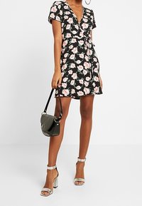 Kvinna klädd i en svart blommig omlottskirt med rosa blommor, som håller en svart handväska och bär silvriga högklackade sandaler.