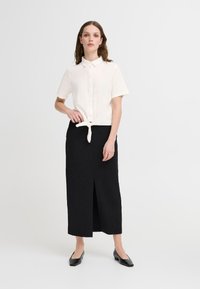 Camicia bianca a maniche corte con colletto e dettaglio con nodo anteriore, abbinata a una gonna nera lunga fino al pavimento con uno spacco frontale. Scarpe piatte nere.
