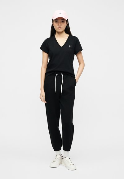 Γυναίκα που φοράει ροζ καπέλο, μαύρο T-shirt με V λαιμόκοψη, μαύρο παντελόνι με κορδόνι στη μέση, λευκές κάλτσες και λευκά αθλητικά παπούτσια, στέκεται μπροστά σε απλό φόντο.