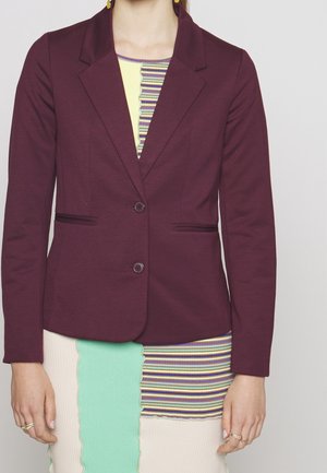 Mujer con blazer entallado color granate sobre una falda de parches a rayas multicolores y lisos, de pie con las manos relajadas a los lados.