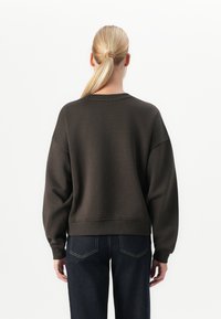 Mujer con cabello rubio en una coleta, vistiendo una sudadera de manga larga marrón oscuro y jeans azul oscuro, de pie de espaldas contra un fondo blanco.