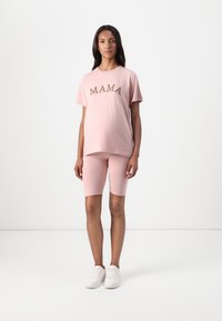 Light pink cotton t-shirt with "MAMA" in brown lettering, paired with matching biker shorts. Model wears white sneakers.