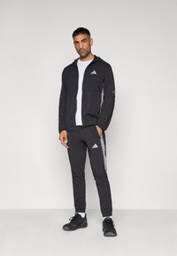 Μαύρο αθλητικό μπουφάν με κουκούλα και μπροστινές τσέπες, σε συνδυασμό με μαύρα τζόγκερ με γκρι λεπτομέρειες, και τα δύο από ελαφρύ υλικό.