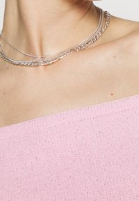 Stříbrný kovový náhrdelník s vrstvami různých stylů řetízků, obsahující hladké a texturované články, nošený na světle růžovém topu.