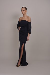 Robe bleu marine à épaules dénudées avec des manches bouffantes, silhouette ajustée et fente haute jusqu'à la cuisse. Tissu lisse avec une légère brillance, associé à des talons dorés.