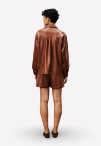 Personne avec des cheveux courts et bouclés, portant une chemise en cuir marron à manches longues et un short assorti, se tenant de dos sur un fond blanc.