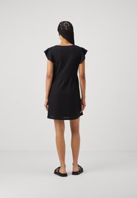 Vestido preto curto com mangas curtas, tecido texturizado, cintura ajustada, detalhes de ilhós na bainha, combinado com sandálias planas pretas.