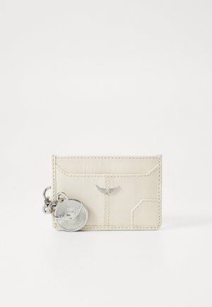 Porte-cartes couleur crème en cuir lisse, avec des accents cousus et un charm en métal argenté présentant un design texturé et circulaire.