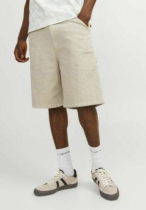 Pantaloni cargo beige con tasche frontali, orli cuciti e vestibilità comoda. Indossati con sneaker beige e calzini bianchi con testo nero.