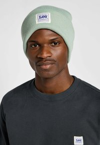 Jasnozielona dzianinowa czapka z metką z niebieskim napisem. Noszona z ciemnoszarym swetrem. Zbliżenie na twarz męskiego modela.