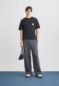 Μαύρο βαμβακερό μπλουζάκι με μπροστινή τσέπη, σε συνδυασμό με γκρι, άνετες φόρμες. Το μοντέλο κρατά μια μικρή μαύρη τσάντα, φοράει ανοιχτόχρωμα αθλητικά παπούτσια.