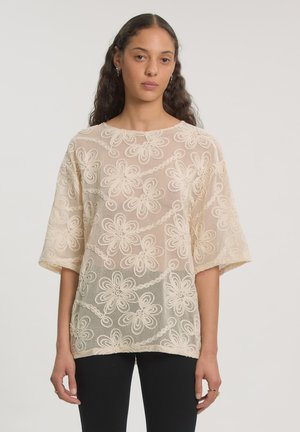 Vrouw die een crèmekleurige kanten top draagt met bloemenpatronen en halflange mouwen, gecombineerd met een zwarte broek, staand tegen een effen achtergrond.