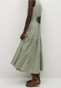Robe maxi fleurie verte avec un corsage ajusté, une jupe évasée, un détail découpé dans le dos et un ourlet volanté. Associée à des sandales marron à lanières.