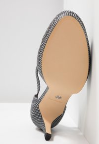Chaussure à talon aiguille noire et blanche à motif, avec un bout pointu, un design ouvert au dos et une semelle beige lisse. Détail du talon visible.
