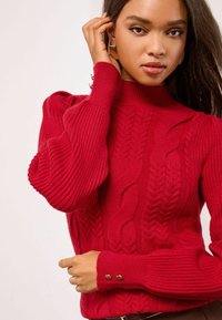 Roter Strickpullover mit Rippen- und Zopfmuster, hohem Kragen und goldenen Knopfakzenten an den Ärmeln. Weicher Stoff und taillierte Passform.