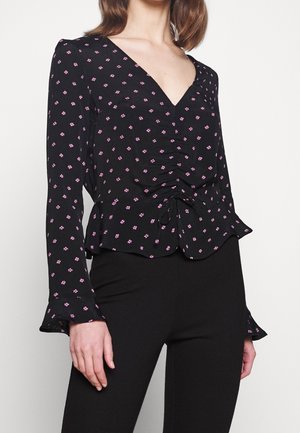 Frau trägt eine schwarze langärmelige V-Ausschnitt-Bluse mit rosa Blumenmuster, geraffter Vorderseite und Rüschenmanschetten, kombiniert mit schwarzen Hosen.