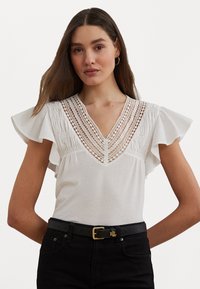 White cotton blouse featuring a V-neckline with lace detailing and ruffled sleeves. Paired with black jeans and a black belt with gold hardware.