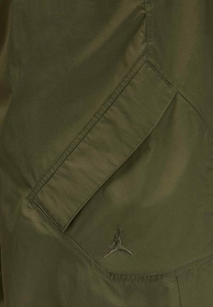 Olive green windbreaker featuring a smooth, lightweight texture, diagonal side pockets, and a black embroidered Jumpman logo.