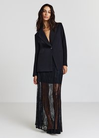 Blazer noir avec col cranté, fermeture à un seul bouton et deux poches avant, associé à une jupe maxi en dentelle noire transparente.